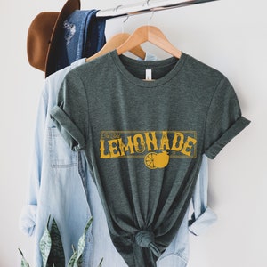 K&ouml;nnte beinhalten: Ein dunkelgr&uuml;nes T-Shirt mit einer gelben Grafik, auf der "Ice Cold Lemonade" mit einer Zitronenscheibe und einem Stern steht.