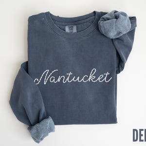 May include: A denim blue sweatshirt with the word "Nantucket" embroidered in white script across the chest. The sleeves are cuffed, and the word "DENIM" is printed in a darker blue at the bottom right.