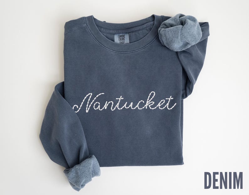 May include: A denim blue sweatshirt with the word "Nantucket" embroidered in white script across the chest. The sleeves are cuffed, and the word "DENIM" is printed in a darker blue at the bottom right.