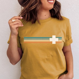 Puede incluir: Una camiseta amarilla mostaza con un gr&aacute;fico de cruz blanca y un dise&ntilde;o a rayas de estilo retro en tonos de azul verdoso, naranja y marr&oacute;n.