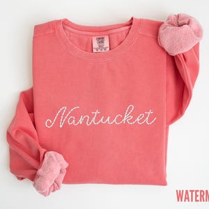 May include: A coral-colored sweatshirt with the word "Nantucket" embroidered in white script across the front. The sleeves are folded, revealing a lighter pink, textured fabric. The word "WATERMELON" is printed in red at the bottom right.