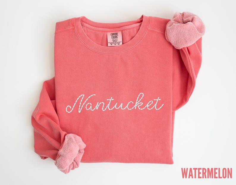 May include: A coral-colored sweatshirt with the word "Nantucket" embroidered in white script across the front. The sleeves are folded, revealing a lighter pink, textured fabric. The word "WATERMELON" is printed in red at the bottom right.