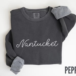 May include: A dark gray sweatshirt with the word "Nantucket" embroidered in white script across the front. The sleeves are cuffed, revealing a gray, fuzzy lining. The word "PEPPER" is printed in a sans-serif font in the bottom right corner.