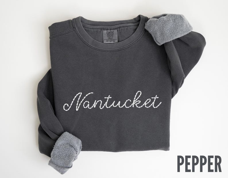 May include: A dark gray sweatshirt with the word "Nantucket" embroidered in white script across the front. The sleeves are cuffed, revealing a gray, fuzzy lining. The word "PEPPER" is printed in a sans-serif font in the bottom right corner.