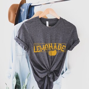 K&ouml;nnte beinhalten: Ein dunkelgraues T-Shirt mit einer gelben Grafik, auf der "Ice Cold Lemonade" mit einer Zitronenscheiben-Illustration steht.