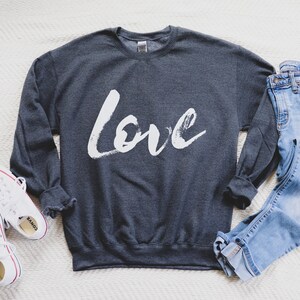 Puede incluir: Una sudadera gris oscuro con la palabra "love" escrita en pintura blanca en la parte delantera. La sudadera est&aacute; colocada sobre una superficie blanca con un par de zapatillas blancas y un par de pantalones vaqueros azules.