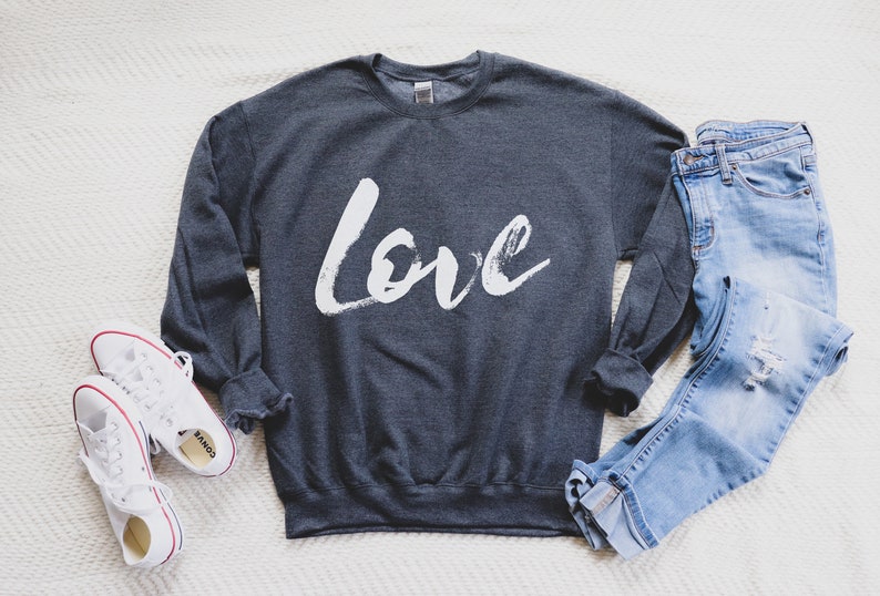 Puede incluir: Una sudadera gris oscuro con la palabra "love" escrita en pintura blanca en la parte delantera. La sudadera est&aacute; colocada sobre una superficie blanca con un par de zapatillas blancas y un par de pantalones vaqueros azules.