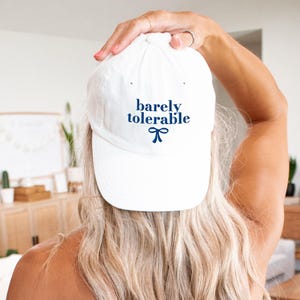 May include: White baseball cap with the text "barely tolerable" in blue lettering and a blue bow.