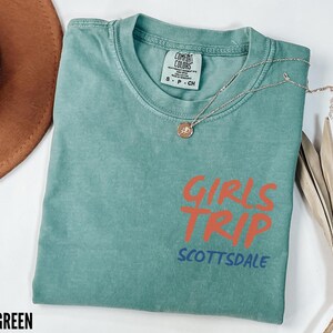 May include: Light green t-shirt with the words "GIRLS TRIP SCOTTSDALE" in orange and blue. The shirt has a crew neck and short sleeves. A gold necklace with a pendant is draped across the shirt.