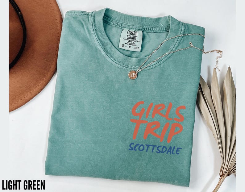 May include: Light green t-shirt with the words "GIRLS TRIP SCOTTSDALE" in orange and blue. The shirt has a crew neck and short sleeves. A gold necklace with a pendant is draped across the shirt.