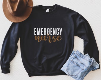 May include: Black sweatshirt with the text "EMERGENCY nurse" in white lettering with a leopard print design. The sweatshirt is laying on a white surface with a brown hat and blue jeans.