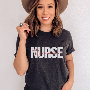 May include: A dark gray t-shirt with the word "NURSE" printed in white letters. The word "trauma" is written in red cursive script underneath the word "NURSE".