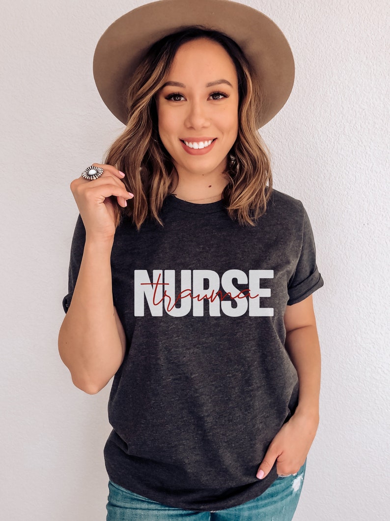 May include: A dark gray t-shirt with the word "NURSE" printed in white letters. The word "trauma" is written in red cursive script underneath the word "NURSE".