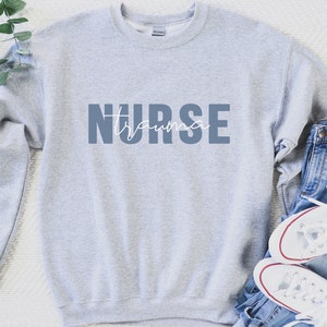 K&ouml;nnte beinhalten: Hellgraues Sweatshirt mit dem Aufdruck "NURSE trauma" in Blau und Wei&szlig;. Das Sweatshirt wird mit blauen Jeans und wei&szlig;en Turnschuhen mit roten Akzenten kombiniert. Ein Zweig mit Gr&uuml;nzeug befindet sich oben links.