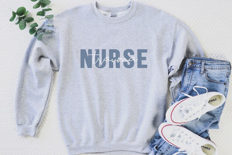K&ouml;nnte beinhalten: Hellgraues Sweatshirt mit dem Aufdruck "NURSE trauma" in Blau und Wei&szlig;. Das Sweatshirt wird mit blauen Jeans und wei&szlig;en Turnschuhen mit roten Akzenten kombiniert. Ein Zweig mit Gr&uuml;nzeug befindet sich oben links.