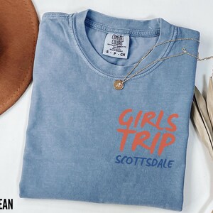 May include: A blue jean colored t-shirt with the words "GIRLS TRIP" in orange and "SCOTTSDALE" in blue. A gold necklace with a pendant is draped across the shirt. A brown hat and dried plant are in the background.