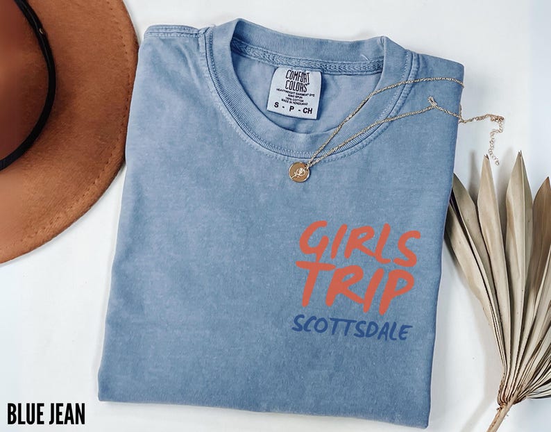 May include: A blue jean colored t-shirt with the words "GIRLS TRIP" in orange and "SCOTTSDALE" in blue. A gold necklace with a pendant is draped across the shirt. A brown hat and dried plant are in the background.