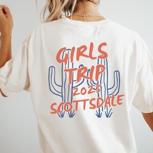 May include: Cream-colored t-shirt with a graphic design on the back. The design features the words "GIRLS TRIP 2025 SCOTTSDALE" in red and blue, along with a cactus illustration. A casual tee for a group vacation.