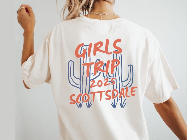 May include: Cream-colored t-shirt with a graphic design on the back. The design features the words "GIRLS TRIP 2025 SCOTTSDALE" in red and blue, along with a cactus illustration. A casual tee for a group vacation.
