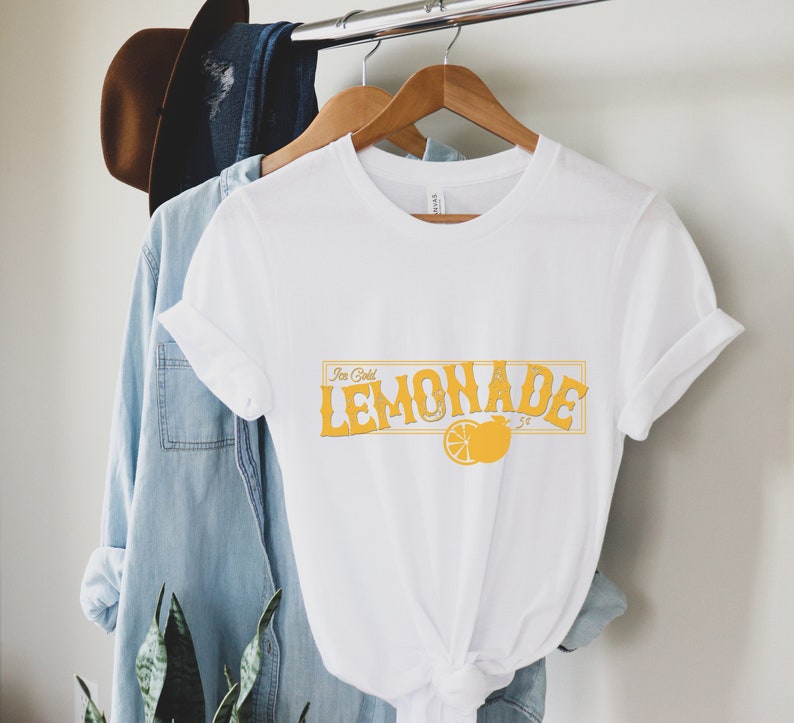 K&ouml;nnte beinhalten: Wei&szlig;es T-Shirt mit einer gelben Grafik, auf der "Ice Cold Lemonade" mit einer Zitronenillustration steht.