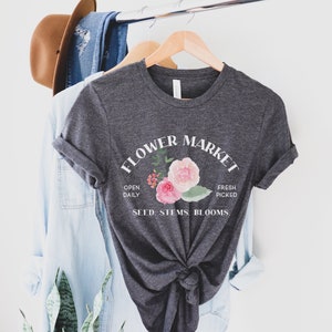 May include: A dark gray t-shirt with a white graphic design that says "Flower Market" with a floral illustration and the text "Open Daily, Fresh Picked, Seed, Stems, Blooms".