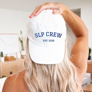 May include: A white baseball cap featuring the text SLP CREW in bold blue letters, with the text EST. 2026 printed in a smaller blue font directly underneath. The cap has a structured crown and a curved brim, presenting a clean and simple design.