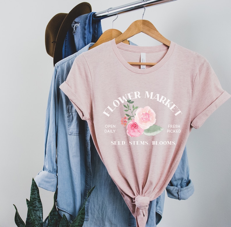 May include: A pink t-shirt with a white graphic that says "Flower Market" with a floral design and the text "Open Daily, Fresh Picked, Seed, Stems, Blooms."