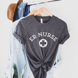 May include: A dark gray t-shirt with the text "ER NURSE" in white lettering and a white plus sign. The shirt is hanging on a wooden hanger.
