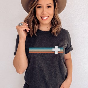 Puede incluir: Una camiseta gris oscuro con una cruz blanca y rayas de colores a trav&eacute;s del pecho. La camiseta est&aacute; hecha de un material suave y c&oacute;modo.