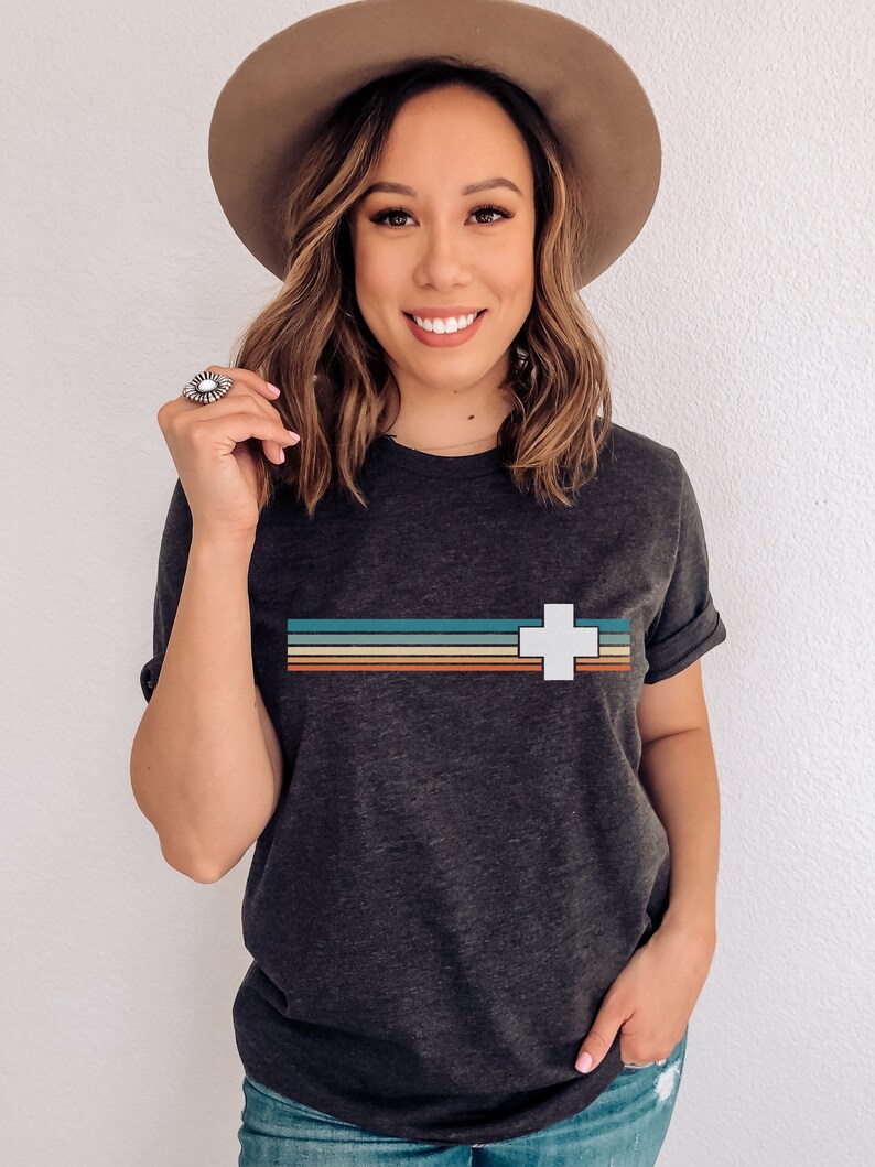 Puede incluir: Una camiseta gris oscuro con una cruz blanca y rayas de colores a trav&eacute;s del pecho. La camiseta est&aacute; hecha de un material suave y c&oacute;modo.
