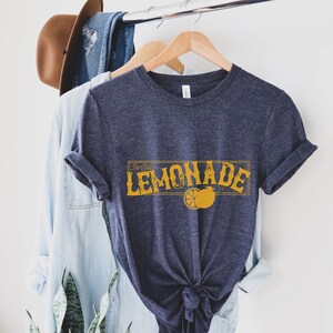 K&ouml;nnte beinhalten: Ein marineblaues T-Shirt mit einer Grafik im Vintage-Stil, auf dem in Gelb "Ice Cold Lemonade" mit einer Illustration einer Zitronenscheibe steht.