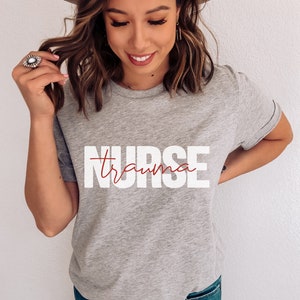 May include: A gray t-shirt with the text "NURSE" in white letters and the word "trauma" written in cursive in red letters.