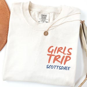 May include: Ivory-colored t-shirt with the words "GIRLS TRIP" in orange and "SCOTTSDALE" in blue. The shirt has a crew neck and short sleeves. A gold necklace with a pendant is draped on the shirt.