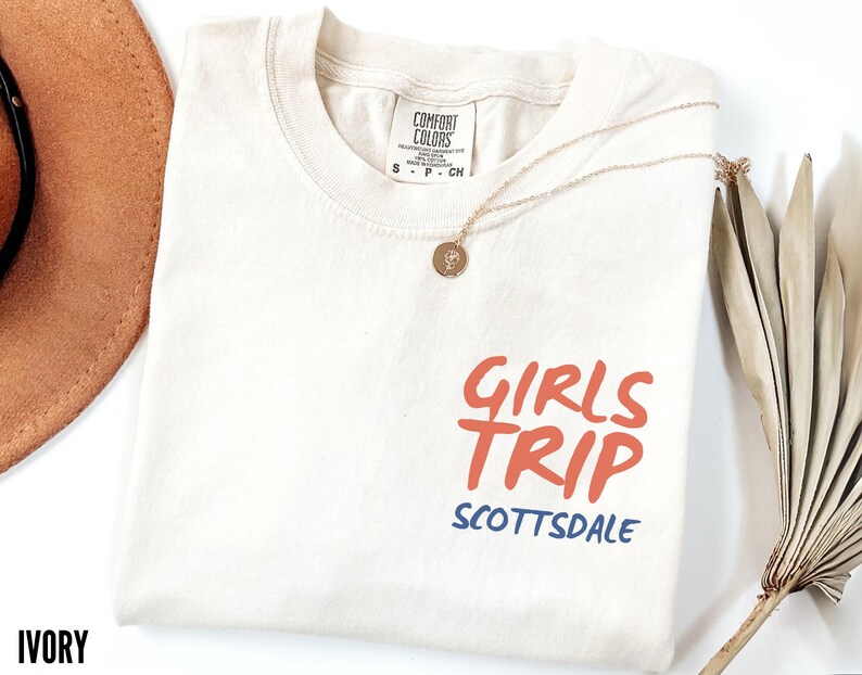 May include: Ivory-colored t-shirt with the words "GIRLS TRIP" in orange and "SCOTTSDALE" in blue. The shirt has a crew neck and short sleeves. A gold necklace with a pendant is draped on the shirt.