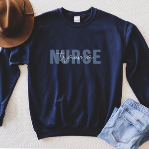 K&ouml;nnte beinhalten: Marineblaues Sweatshirt mit dem Schriftzug "NURSE trauma" in Hellblau und Wei&szlig;. Ein brauner Hut und eine blaue Jeans liegen neben dem Sweatshirt. Das Sweatshirt hat einen Rundhalsausschnitt.