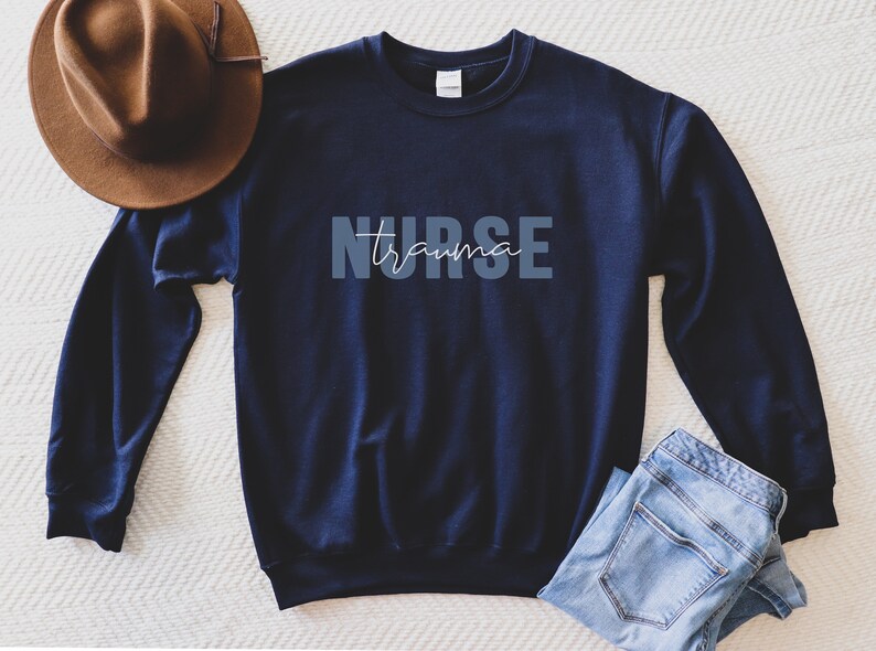 K&ouml;nnte beinhalten: Marineblaues Sweatshirt mit dem Schriftzug "NURSE trauma" in Hellblau und Wei&szlig;. Ein brauner Hut und eine blaue Jeans liegen neben dem Sweatshirt. Das Sweatshirt hat einen Rundhalsausschnitt.