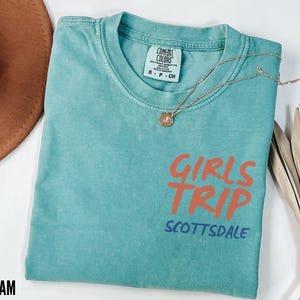May include: Seafoam green t-shirt with the words "GIRLS TRIP SCOTTSDALE" in orange and blue. A gold necklace with a pendant is draped across the shirt. A brown hat is visible in the background.