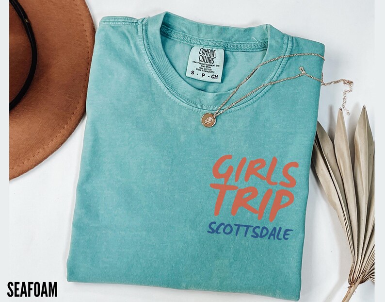 May include: Seafoam green t-shirt with the words "GIRLS TRIP SCOTTSDALE" in orange and blue. A gold necklace with a pendant is draped across the shirt. A brown hat is visible in the background.