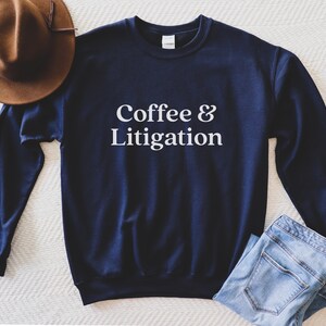 Op de afbeelding: Een marineblauw sweatshirt met de tekst "Coffee & Litigation" in wit gedrukt op de voorkant.