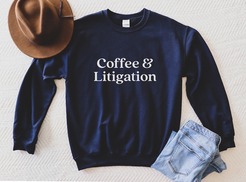 Op de afbeelding: Een marineblauw sweatshirt met de tekst "Coffee & Litigation" in wit gedrukt op de voorkant.