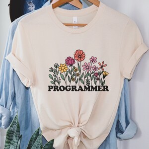 May include: A cream colored t-shirt with a graphic of colorful flowers and the word "PROGRAMMER" in black letters.