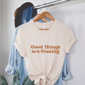 Puede incluir: Una camiseta de color crema con el texto "Good Things are Coming" en una fuente naranja retro.