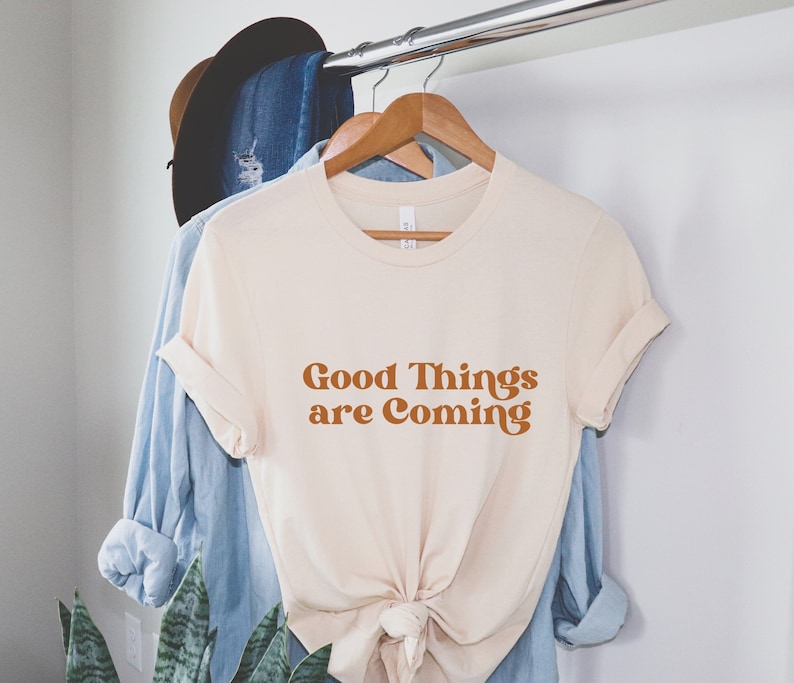 Puede incluir: Una camiseta de color crema con el texto "Good Things are Coming" en una fuente naranja retro.