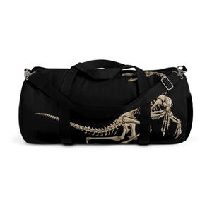Puede incluir: Bolsa de deporte negra con un diseño de esqueleto de dinosaurio beige. La bolsa tiene una correa para el hombro y dos asas. El esqueleto del dinosaurio está representado con detalle, mostrando costillas, columna vertebral y cráneo.