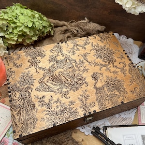 May include: A rectangular wooden box with a hinged lid featuring a dark brown printed toile pattern of figures in period clothing surrounded by floral motifs. The box has visible finger joints at the corners and a small metal latch on the front.