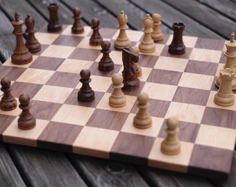 May include: A wooden chessboard with black and white squares. The board is set up with chess pieces in the starting position. The pieces are made of wood and are painted in brown and white.