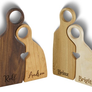May include: Two wooden cutting boards shaped like people, with a heart cutout where they touch. The boards are personalized with the names "Rolf", "Andrea", "Heinz", and "Brigitte".
