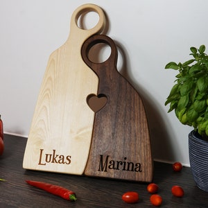 May include: Two wooden cutting boards, one light wood and one dark wood, are joined together in the shape of a heart. The boards are engraved with the names "Lukas" and "Marina".