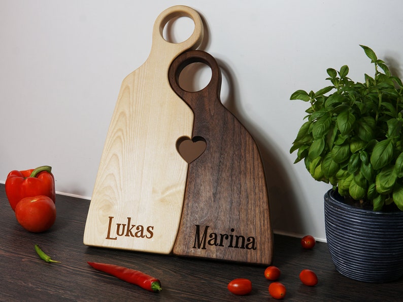 May include: Two wooden cutting boards, one light wood and one dark wood, are joined together in the shape of a heart. The boards are engraved with the names "Lukas" and "Marina".
