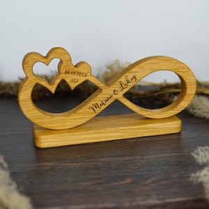 May include: A wooden infinity symbol with two hearts and the engraved text "Marina & Lukas" and "16.08.2015". The wooden sculpture is light brown and sits on a rectangular base. A romantic wedding or anniversary gift.